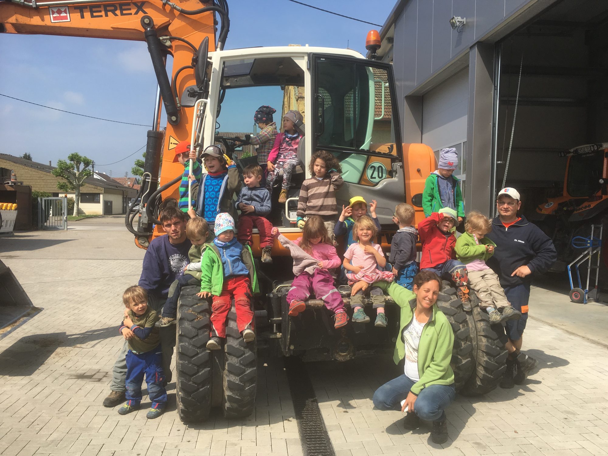 Kindergartengruppe sitzt mit Erzieherinnen und Bauhof-Mitarbeitern auf einem orangen Bagger