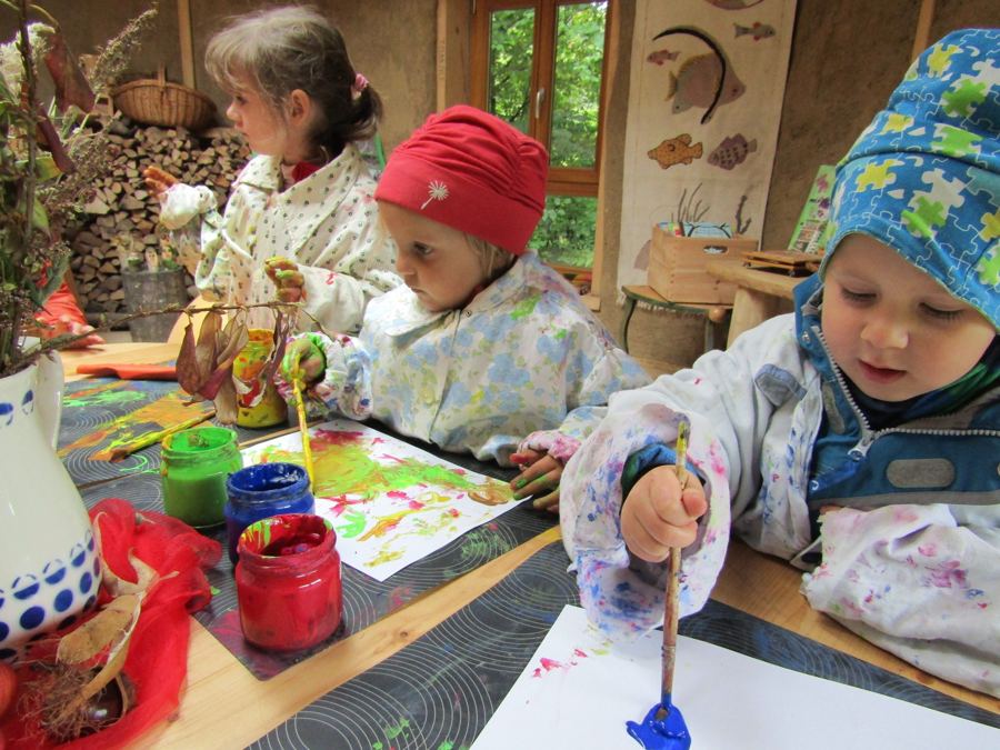 Kinder in Malkitteln malen mit Pinsel und Fingerfarben am Tisch